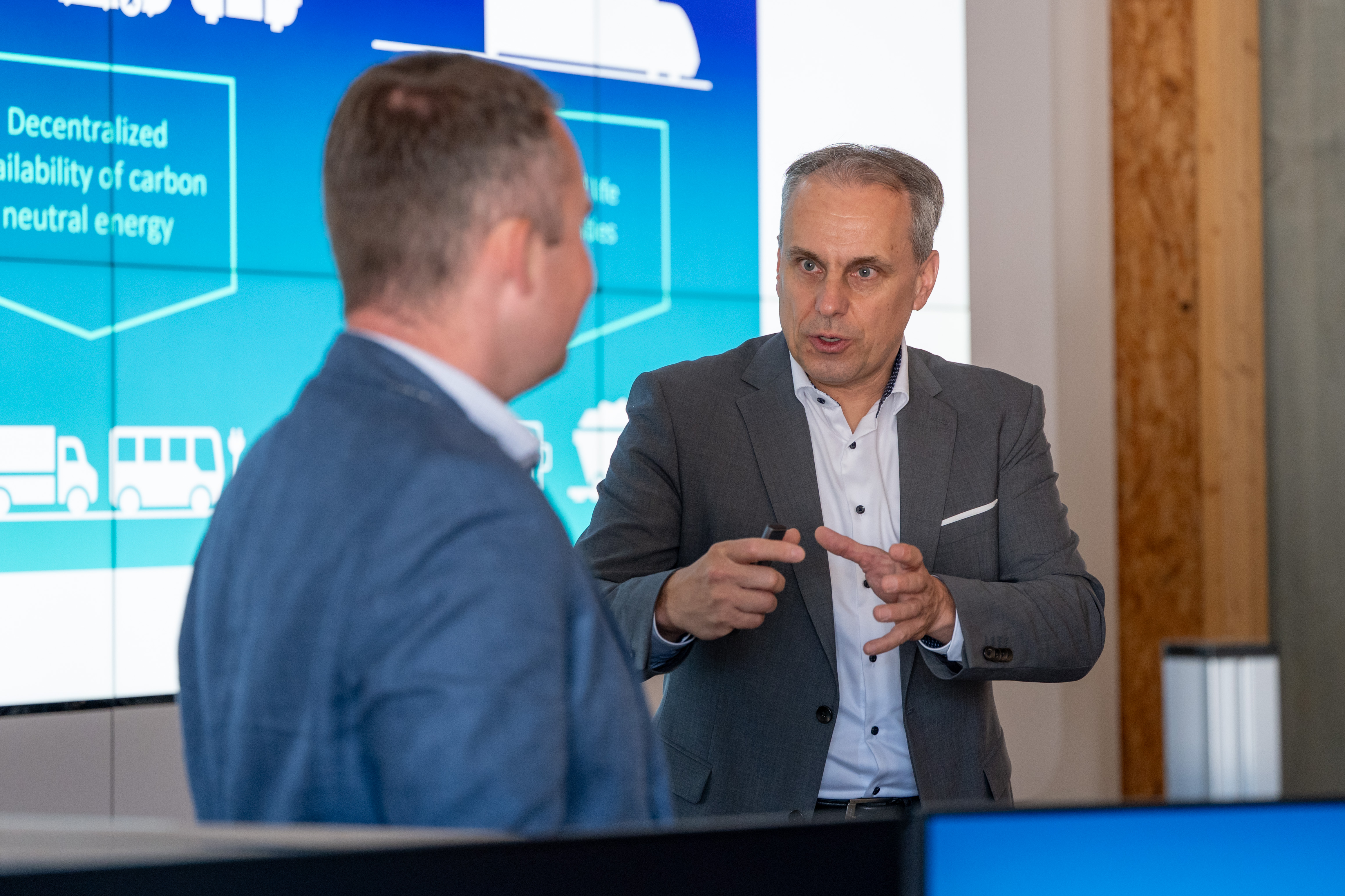 Zwei Männer in Sakkos führen ein Gespräch vor einer Präsentationswand zu neutraler Energie.