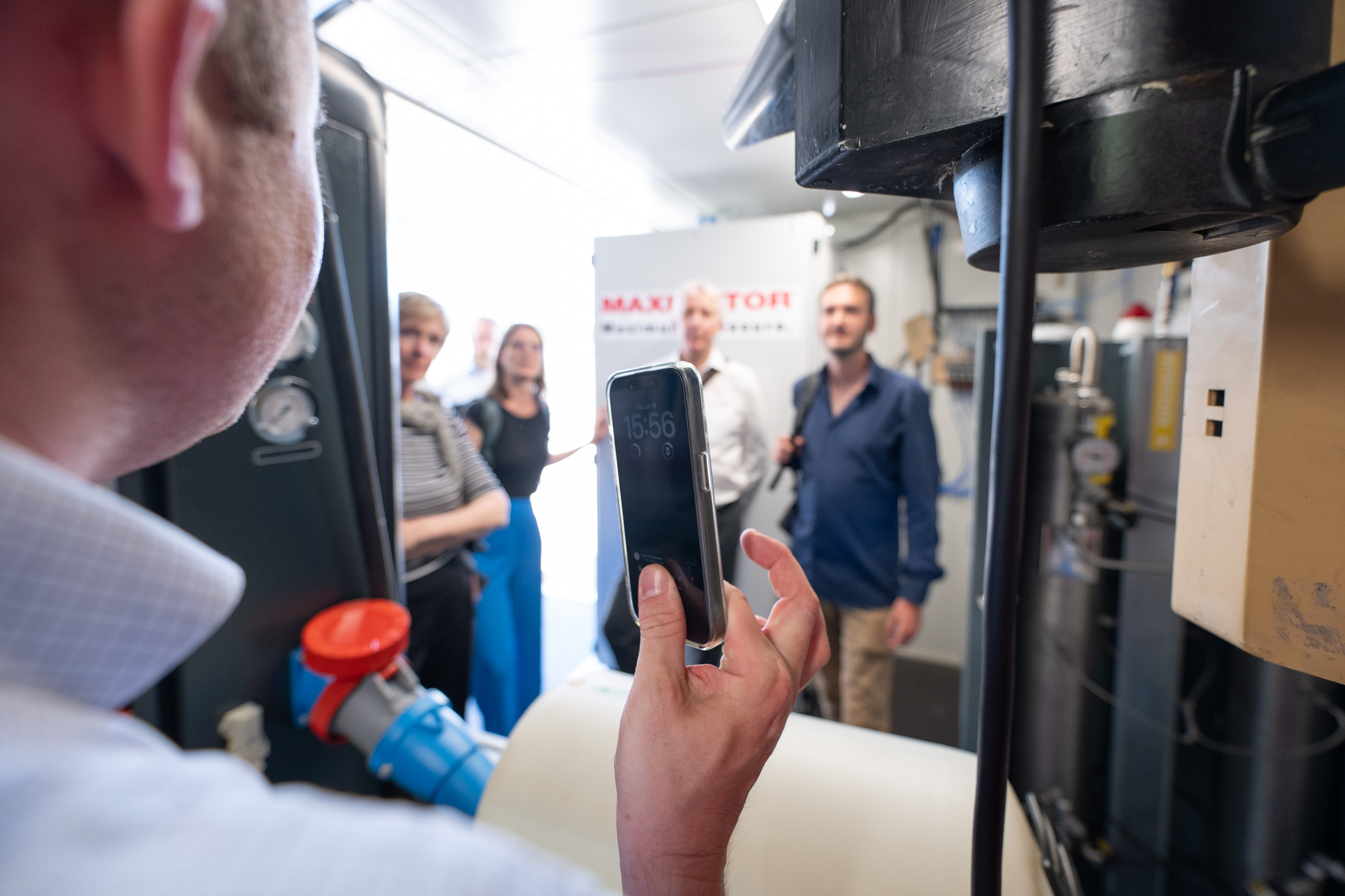 Mann fotografiert eine Gruppe von Menschen in einer technischen Anlage mit dem Smartphone.