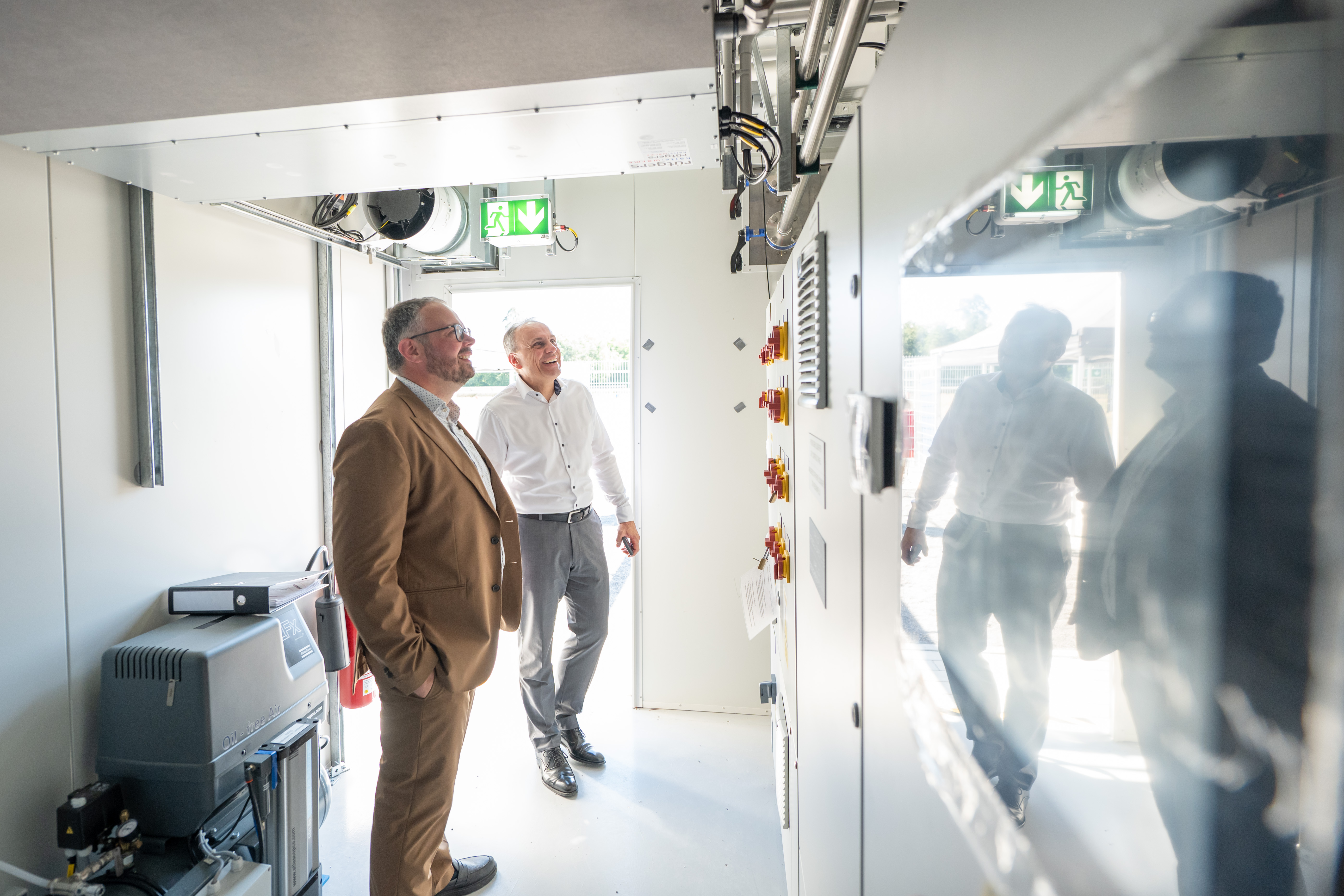 Zwei Männer in Business-Kleidung betrachten eine technische Anlage in einem Technikraum.